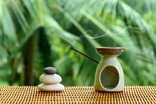 Lámpara de aroma con varita de incienso y piedras zen sobre una mesa de ratán sobre un fondo de naturaleza verde borrosa