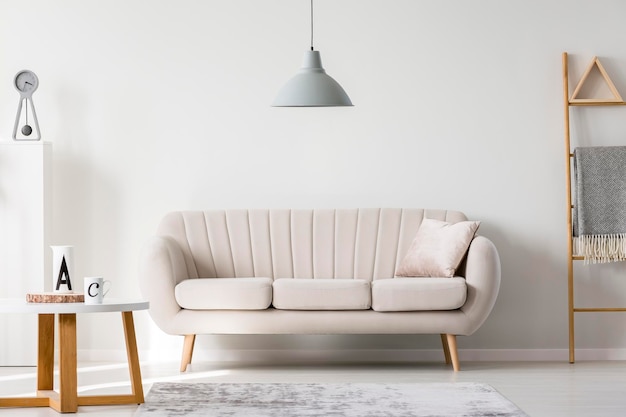 Lámpara gris encima de la almohada en el sofá blanco junto a la mesa de centro de madera con espacio para copiar tazas en la pared vacía