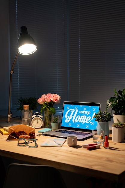 Lámpara sobre mesa de madera con flores, plantas domésticas, laptop, snack, bebida, despertador, productos cosméticos, anteojos y cuaderno en cuarto oscuro