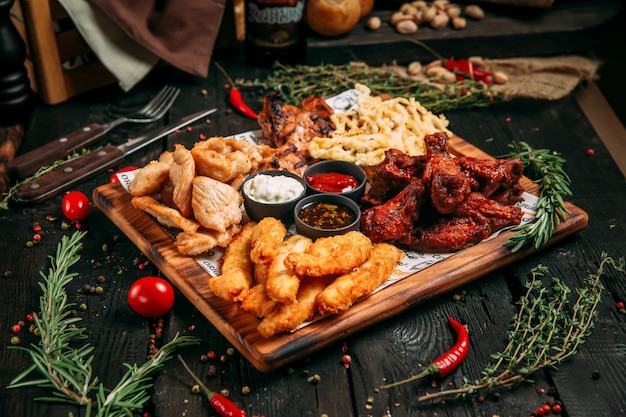 Lanche de cerveja com pedaços de peito de frango, asas, queijo e peixe com molhos na placa de madeira sobre o fundo preto de madeira