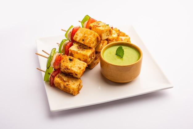 Lanche inicial Paneer Tikka com pau no prato com chutney verde isolado no branco. Prato da culinária indiana com requeijão grelhado com vegetais e temperos