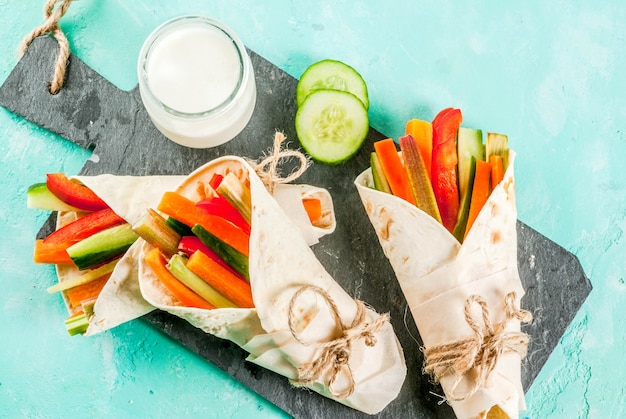 Lanche saudável do verão, sanduíche de tortilla de estilo mexicano envolve varas coloridas de legumes frescos variadas (pepino e pimenta de aipo com ruibarbo e aipo) com molho de iogurte, mergulho - fundo azul claro