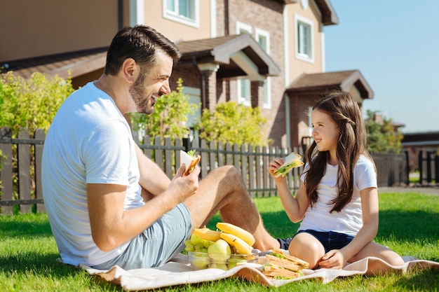 Lanches deliciosos. Agradável jovem pai e sua linda filha sentados na grama comendo sanduíches enquanto sorriem um para o outro