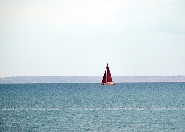 Landschaft mit einer Yacht mit hohen roten Segeln, die in ruhigem Wasser am hügeligen Ufer ins Meer schwebt