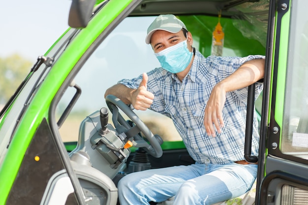 Landwirt fährt Traktor im Maisfeld, der ein Masken-Coronavirus-Konzept trägt