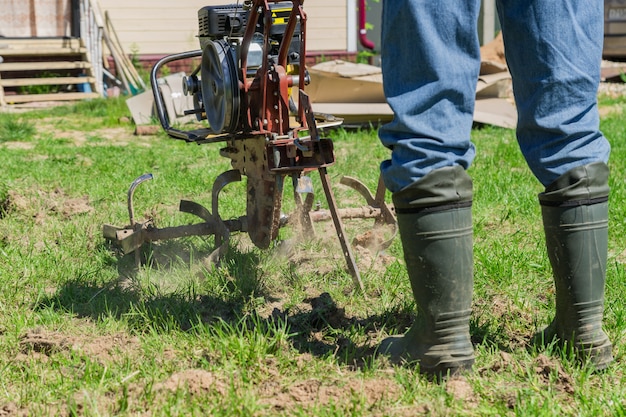 Landwirt in Gummistiefeln und Blue Jeans pflügt einen Boden mit kleinem Motortraktor.