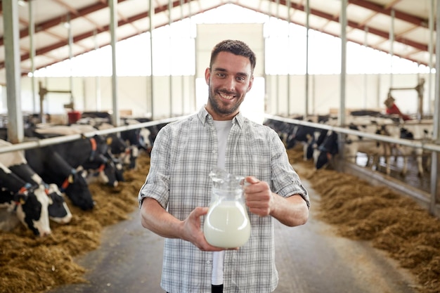 Landwirtschaft, Industrie, Landwirtschaft, Menschen und Viehzucht Konzept - glücklicher lächelnder junger Mann oder Bauer mit Kuhmilch in einem Krug im Kuhstall auf einer Milchfarm