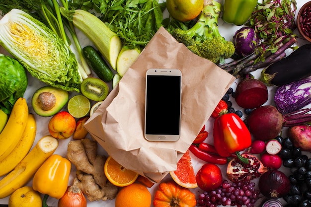 Landwirtschaftliche Produkte, Smartphone auf der Papiertüte - Lieferkonzept, Null-Abfall-Paket. Lebensmittel vom lokalen Markt mit Gemüse und Früchten.