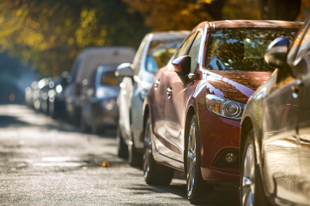 Lange Reihe verschiedener glänzender Autos und Lieferwagen, die am sonnigen Herbsttag am leeren Straßenrand geparkt wurden. Moderner Stadtlebensstil, Fahrzeugparkproblemkonzept.