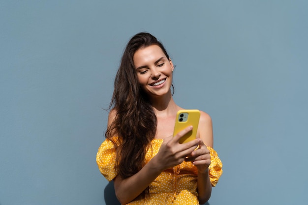Langes dunkles Haar der hübschen Frau im hellen gelben Sommerkleid, das auf blauem Wandhintergrund-Tageslicht aufwirft und chattet und Nachrichten auf dem Handy liest