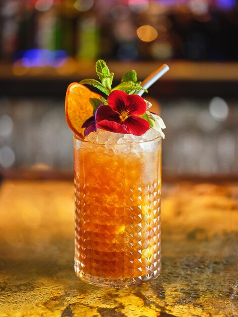 Langes Getränk-Cocktail mit Eiswürfeln, geschmückt mit Blumen und getrockneten Orangenflecken, serviert in einer modernen Bar auf verschwommenem Hintergrund