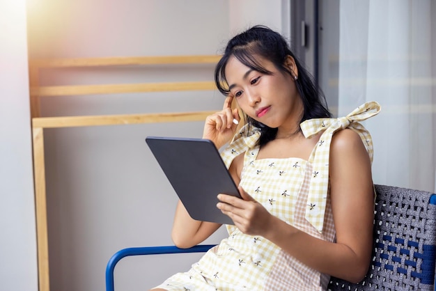 Langhaarige asiatische Frau in einem gelben Kleid liegt glücklich auf ihrem Tablet auf dem bequemen Sofa Online-Shopping-Social-Tech-Kommunikation