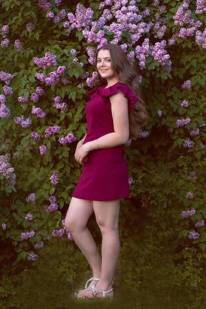 langhaarige braunhaarige frau in einem lila kleid steht vor dem hintergrund eines fliederbusches