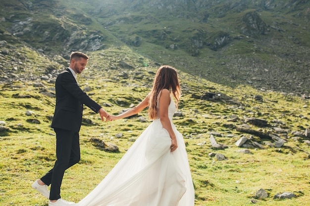 Langhaarige Braut im Hochzeitskleid und Bräutigam, die Händchen halten und bei Sonnenuntergang am Berghang spazieren gehen