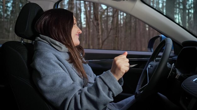 Langhaarige brünette Frau sitzt am Lenkrad eines ausländischen Autos und hört Musik tanzen und lächelt glücklich in der Nähe des Waldes geparkt