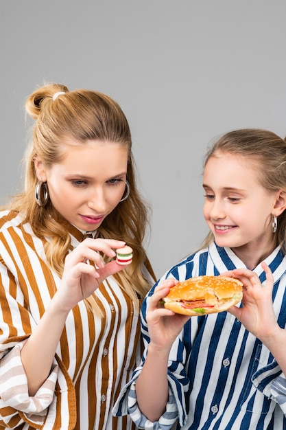 Langhaarige erwachsene Frau, die ihren kleinen gefälschten Burger beobachtet, während die kleine Schwester die Vollversion hält holding