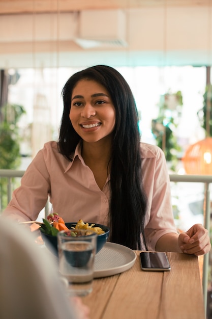 Langhaarige hübsche Dame, die lächelt, während sie ihr Mittagessen in einem schönen Café genießt?