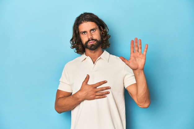 Langhaariger Mann in einem weißen Polo-Blauen Studio, der einen Eid leistet und seine Hand auf die Brust legt