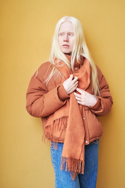 Langhaariges Albino-Mädchen in brauner Jacke und Schal, das an der gelben Wand steht