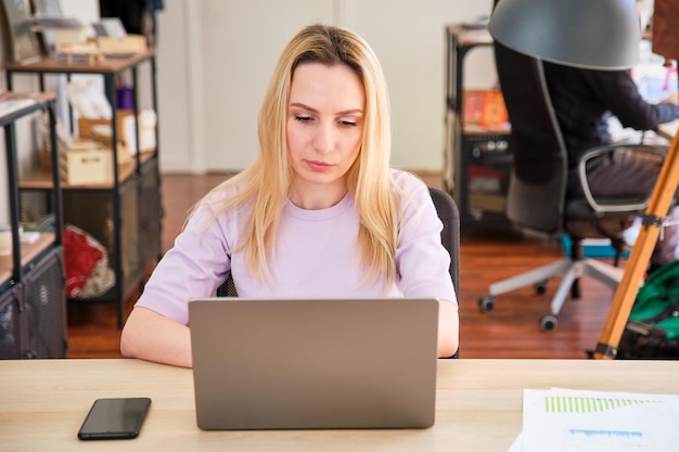 Langhaariges blondes hübsches Mädchen, das mit einem Laptop in einem Büro arbeitet