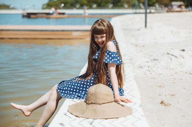 Langhaariges kleines Mädchen in einem Strandhut sitzt am Strand und betrachtet den Seesommer