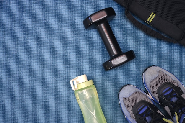 Langhantel eine gelbe Wasserflasche, eine schwarze Tasche und ein Paar Schuhe auf einer blauen Matte für den Trainingshintergrund