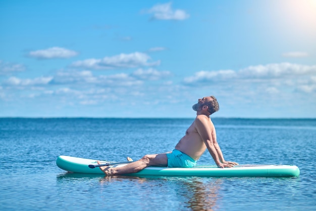 Langsames Leben. Grauhaariger bärtiger Mann, der seitlich zur Kamera sitzt und an einem sonnigen Tag auf dem Paddelbrett auf dem Wasser meditiert