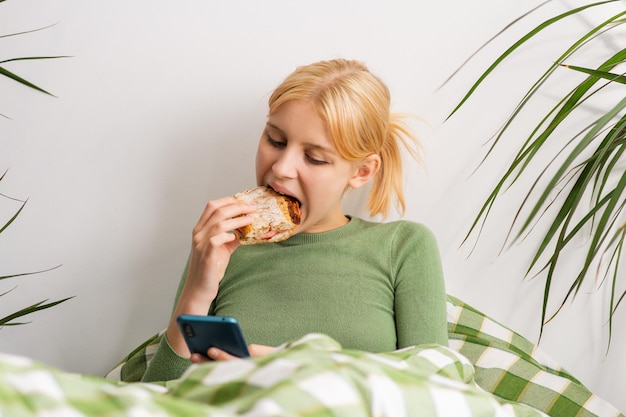 Foto languideciendo en la cama, una niña se enfrenta a la decadencia mental pegada a su teléfono y mordisqueando una tostada durante todo el día.