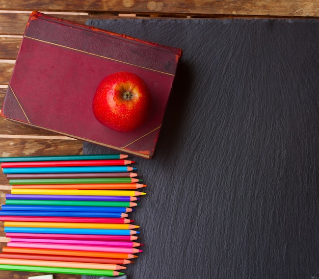 Lápices de colores con libro antiguo y manzana roja en pizarra