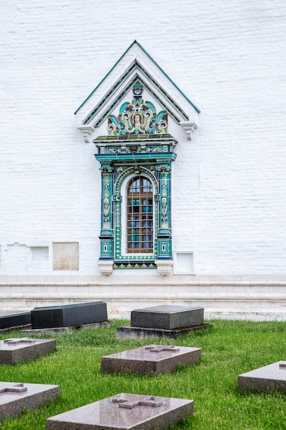 Lápides de mármore em um gramado verde em um cemitério perto da parede de tijolos brancos do templo com uma bela janela esculpida com barras verticais