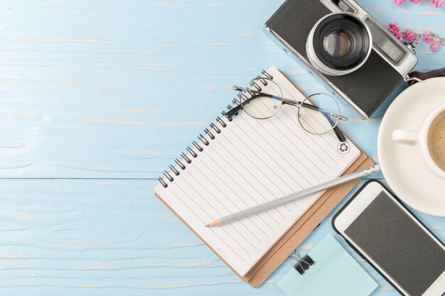 Lápiz en el cuaderno en blanco con cámara vintage y teléfono inteligente sobre fondo de madera vieja, viajes y Hol