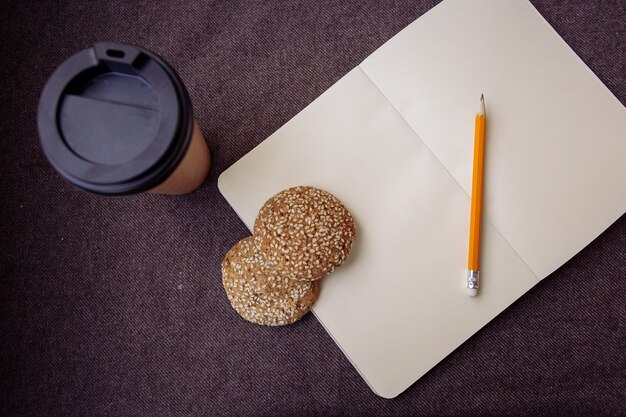 Lápiz, cuaderno, galletas y taza de café de papel sobre un fondo de tela marrón