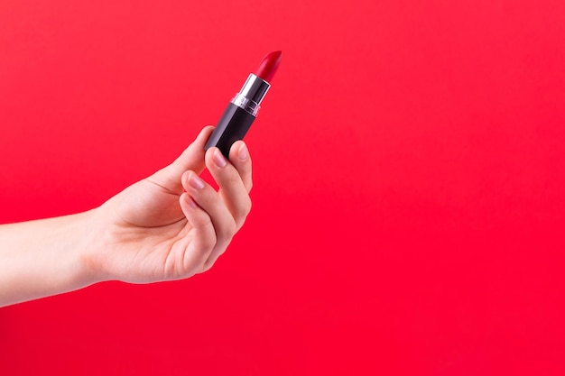 Lápiz labial rojo en un tubo negro sostiene la mano femenina sobre fondo rojo Producto cosmético para una belleza brillante