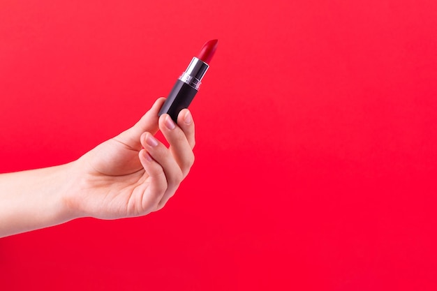 Lápiz labial rojo en un tubo negro sostiene la mano femenina sobre fondo rojo Producto cosmético para una belleza brillante