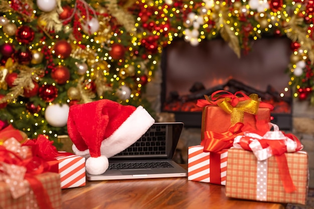 Laptop auf dem Tisch mit Weihnachtsmütze und Weihnachtsgeschenken vor dem Kamin und einem geschmückten Weihnachtsbaum. Online-Konzept