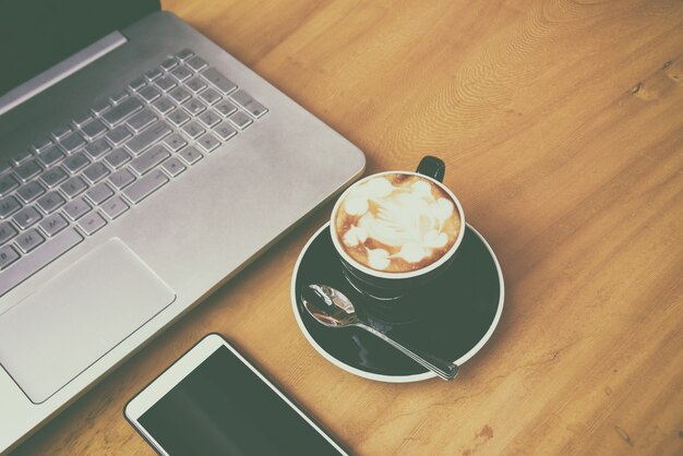 Foto laptop com o telefone do smarth na mesa de madeira no quarto do negócio. uma xícara de café no intervalo de café (efeito de grão de filme)