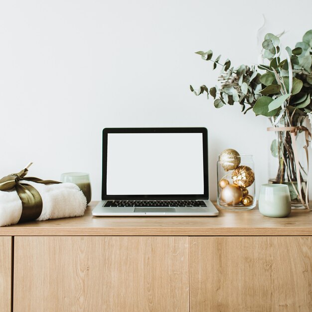 Laptop com tela na área de trabalho de madeira decorada com buquê de eucalipto, caixa de presente, velas na superfície branca.