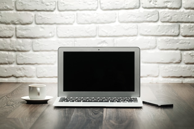 Laptop com tela preta em branco em uma mesa de madeira contra a parede de tijolos brancos