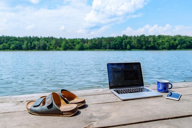 Laptop com xícara e telefone, um rio de doca de madeira no fundo do horário de verão, trabalha com você