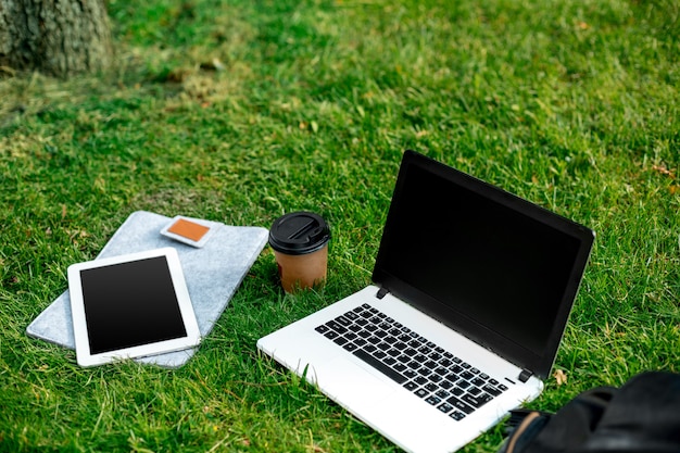 Laptop-Computer auf grünem Gras mit Kaffeetasse, Tasche und Tablet im Park im Freien. Platz kopieren. Stillleben