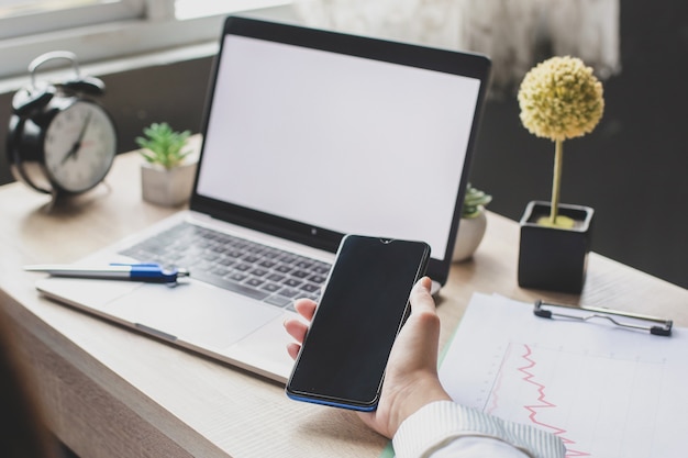 Laptop de tela em branco com a mão segurando um telefone inteligente na mesa do escritório