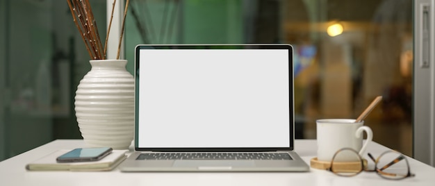 Laptop de tela em branco na mesa de trabalho com copo, smartphone, suprimentos e decoração na sala de escritório de partição de vidro