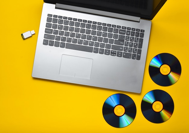 Laptop, drives de CD, pen drive USB em um fundo amarelo. Mídia digital moderna e desatualizada.