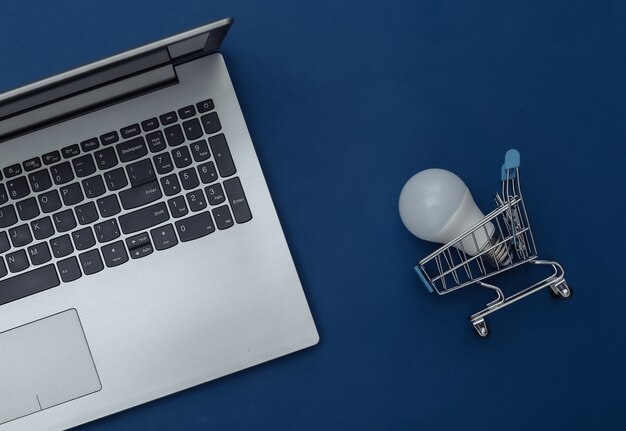 Laptop e carrinho de compras com lâmpada LED em fundo azul clássico. Economize energia. Cor 2020. Vista superior