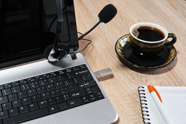 Laptop, fones de ouvido com microfone, notebook com caneta e xícara de café em cima da mesa close up