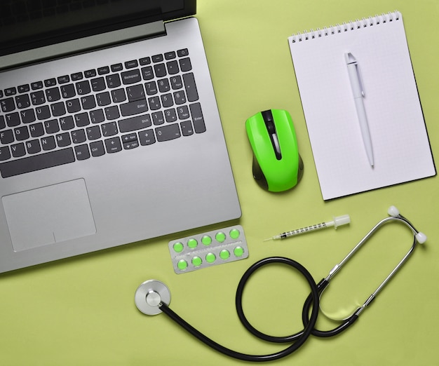 Laptop, kabellose Maus, Notizbuch, Stethoskop, Pillen, Spritze auf einem grünen Pastellhintergrund