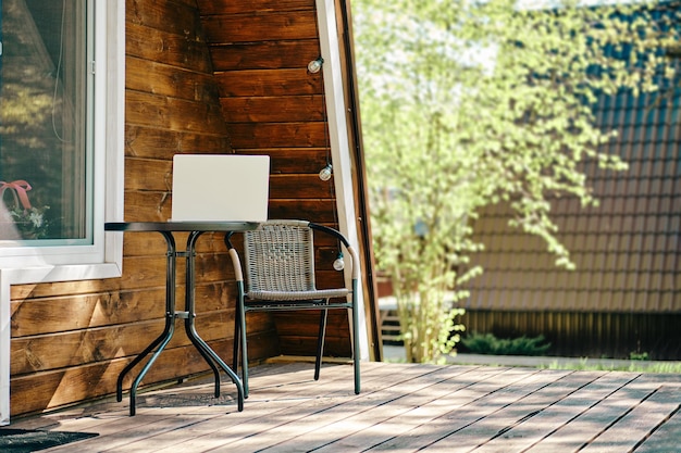 Laptop en la mesa de café cerca de la silla de mimbre en la terraza de la cabaña de madera Concepto de trabajo remoto y freelance