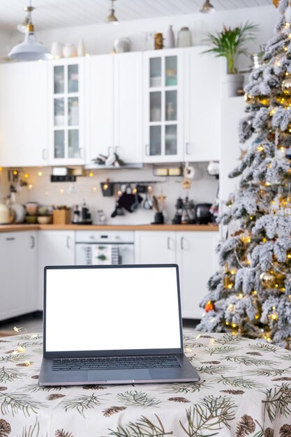 Laptop mit einem weißen Bildschirm im gemütlichen, weiß dekorierten Weihnachtsküchen mit Feenlichtern