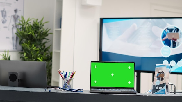 Laptop mit Greenscreen-Display am Schreibtisch auf leerer Registrierungstheke im Wartebereich der Rezeption. Isolierter Copyspace auf dem Bildschirm in der Krankenhauslobby, leeres Chroma-Key-Mockup.