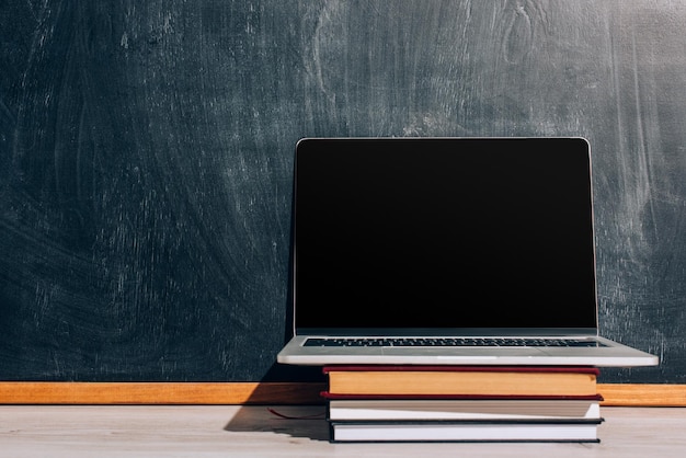 Laptop mit leerem Bildschirm auf einem Stapel Lehrbücher in der Nähe einer schwarzen Tafel